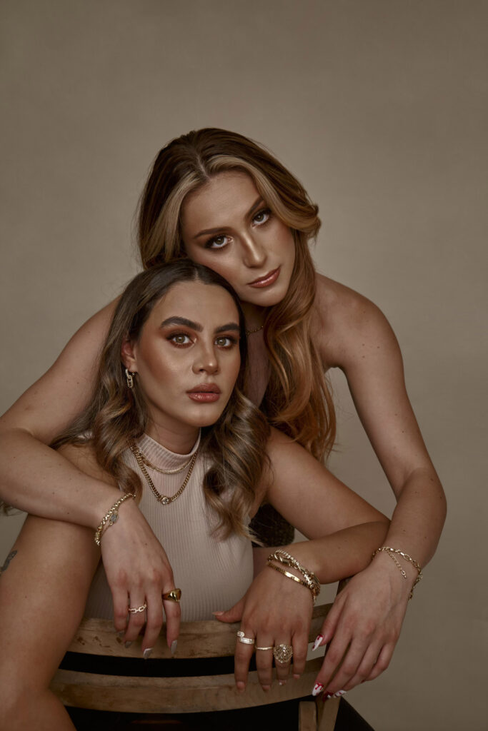 Clarisa and Valeria two women with striking features pose together against a muted background. Clarisa with wavy brown hair wears a beige top and gold jewelry while Valeria has long blonde hair and is adorned in a black outfit with gold accessories.